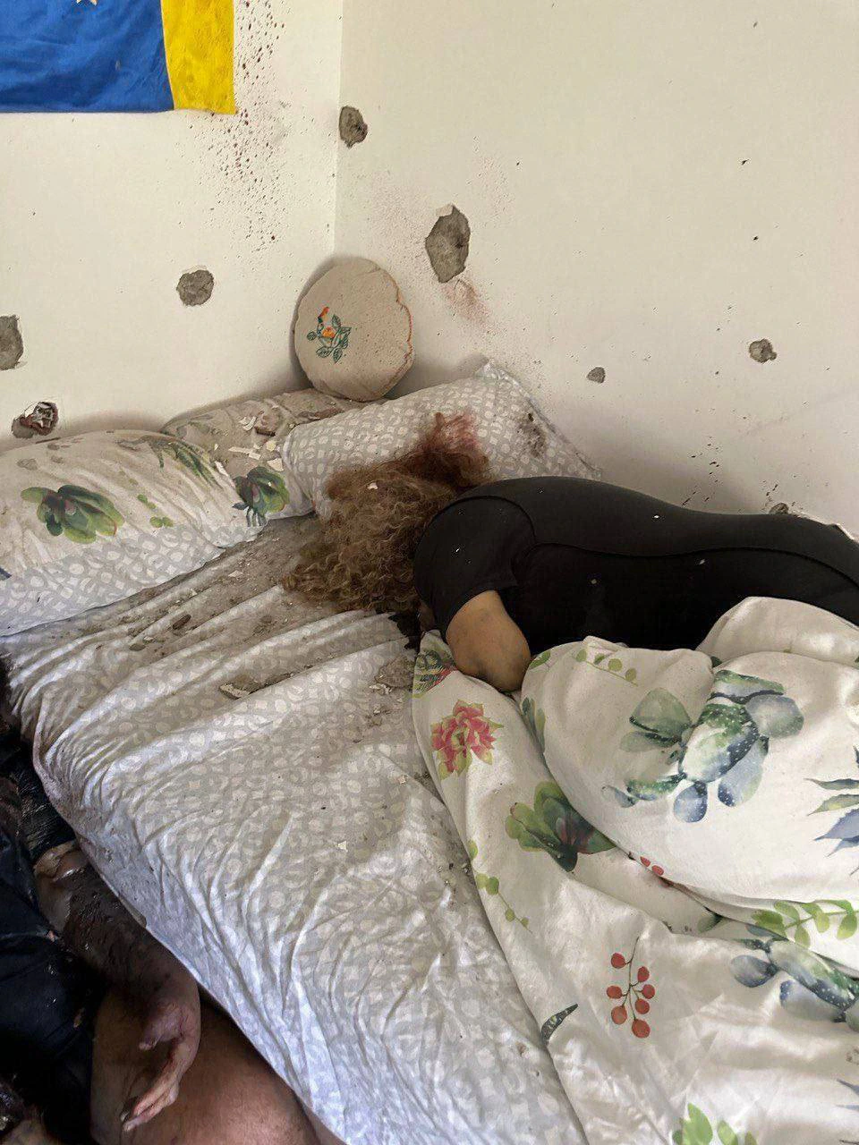 גופת אישה שרועה על מיטה בחדר שינה, ובקירות החדר סביבה חורי ירי