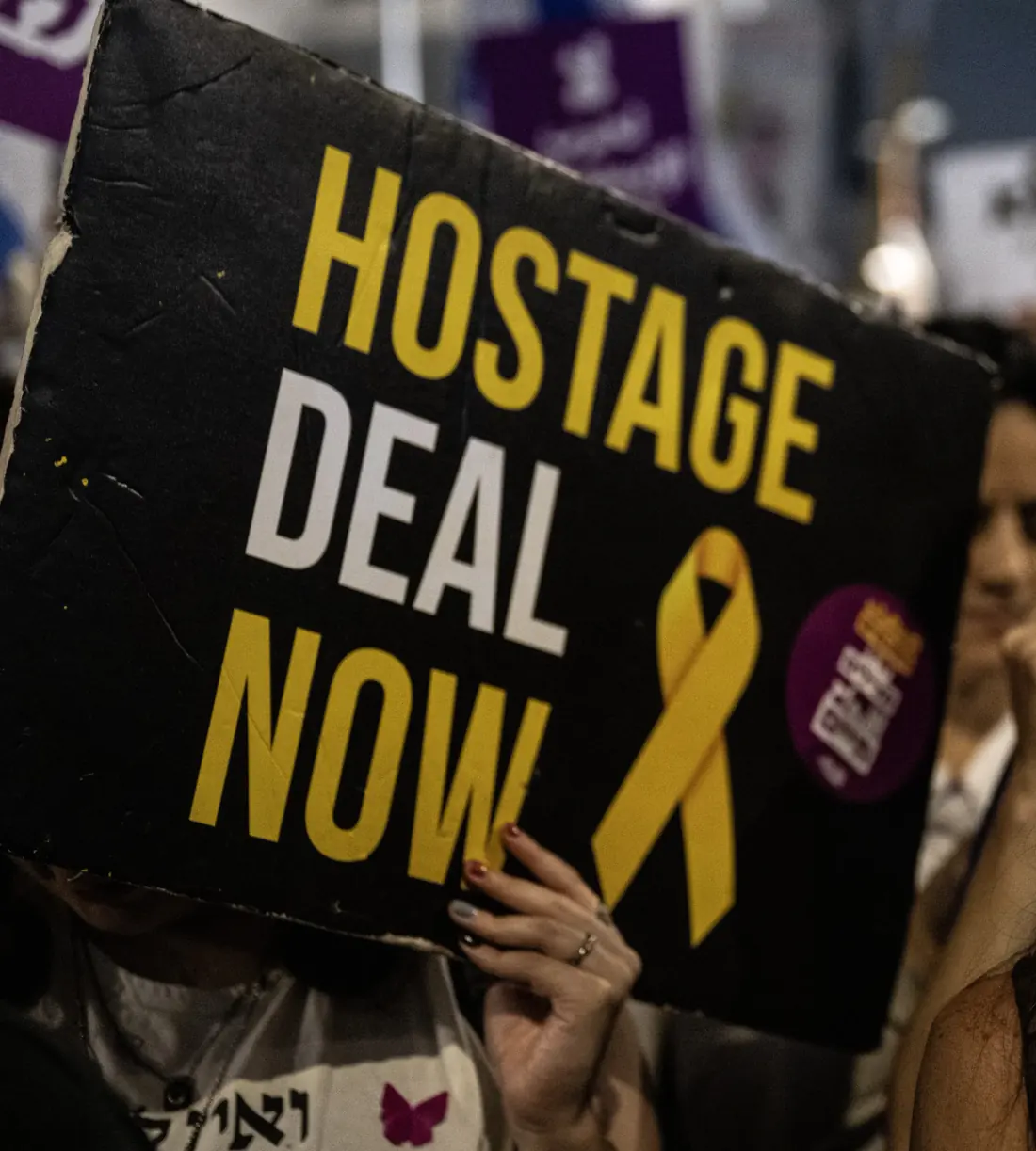 מפגינה מחזיקה שלט שחור ועליו "HOSTAGE DEAL NOW" וסרט צהוב, בהפגנה ערב