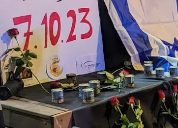 שולחן זיכרון ועליו נרות נשמה ושושנים אדומות, לצד שלט "7.10.23" ודגל ישראל