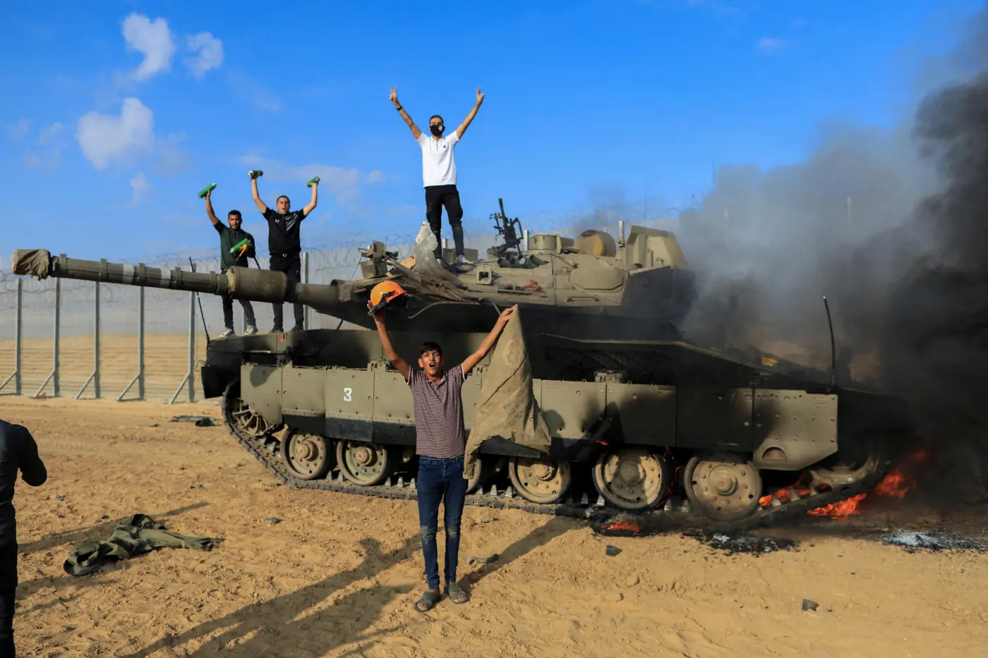 גברים חוגגים על טנק צבאי בוער ולצידו, ליד גדר הגבול, ועשן שחור מיתמר ממנו