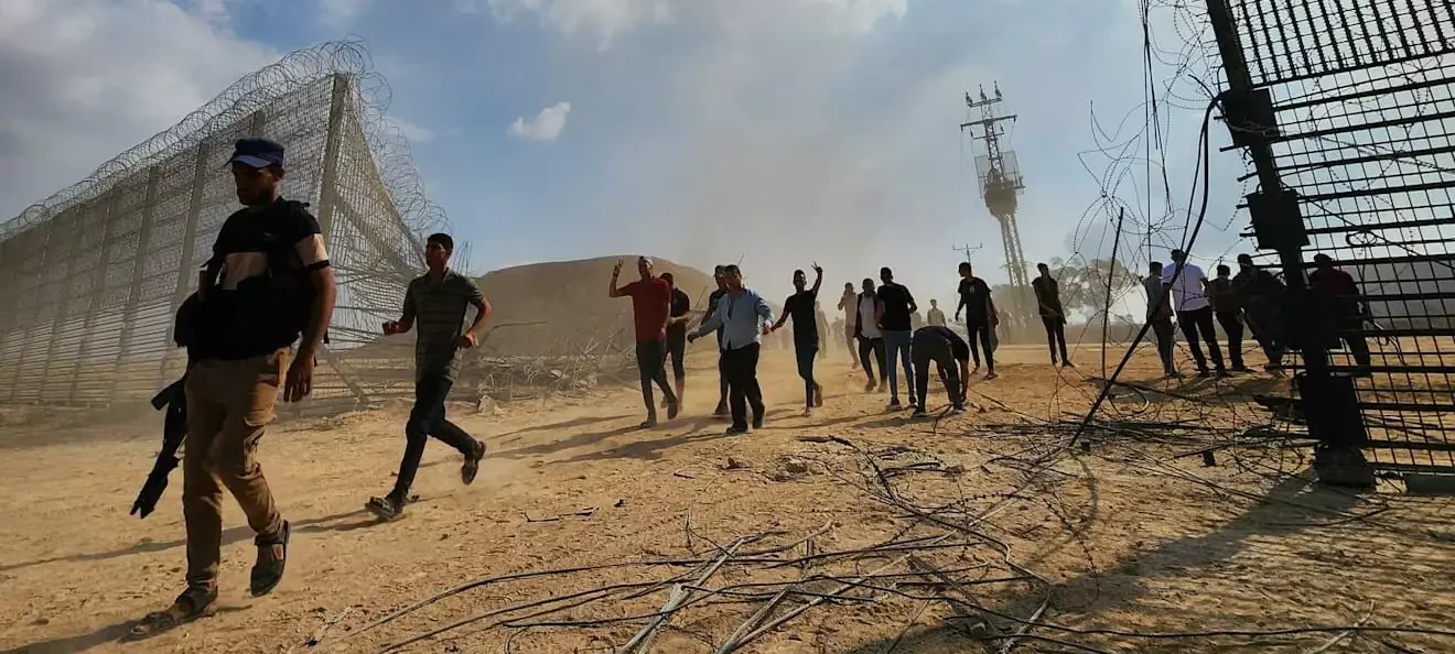 אנשים חוצים ברגל גדר גבול פרוצה בשטח חולי, אחד מהם נושא רובה