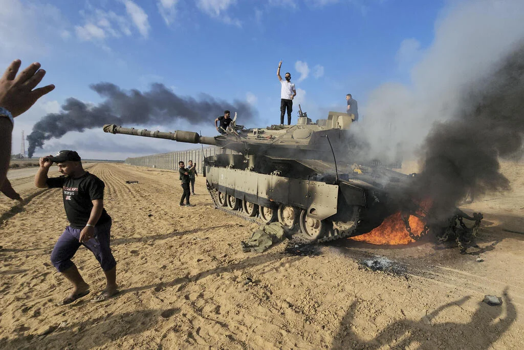 גברים עומדים על טנק צבאי בוער בשטח חולי ליד גדר הגבול, ועשן שחור עולה